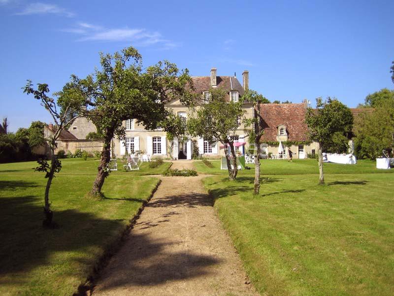 Allée en gravier menant à une maison de campagne entourée d'arbres et d'une pelouse sous un ciel bleu.
