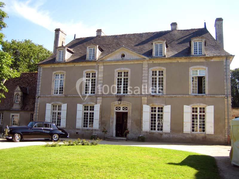 Façade d'une grande maison ancienne avec des volets blancs, une pelouse devant et une voiture ancienne garée.