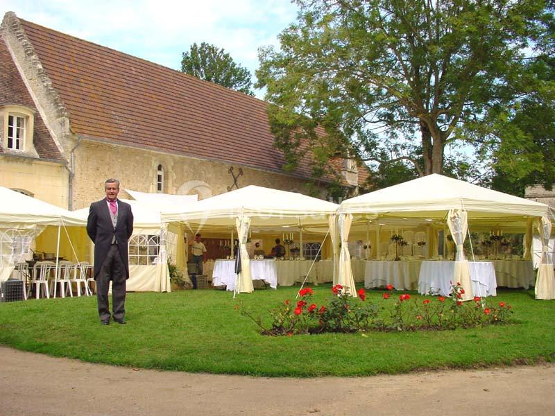 Homme debout devant des tentes blanches installées dans un jardin, près d'un bâtiment ancien avec un toit en tuiles.