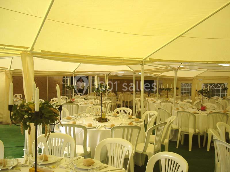 Intérieur d'une tente de réception avec des tables rondes dressées, chaises blanches et décorations florales.