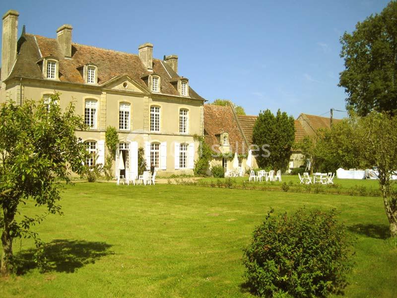 Manoir ancien en pierre avec toit en tuiles, entouré d'un grand jardin verdoyant et meublé de tables et chaises blanches.