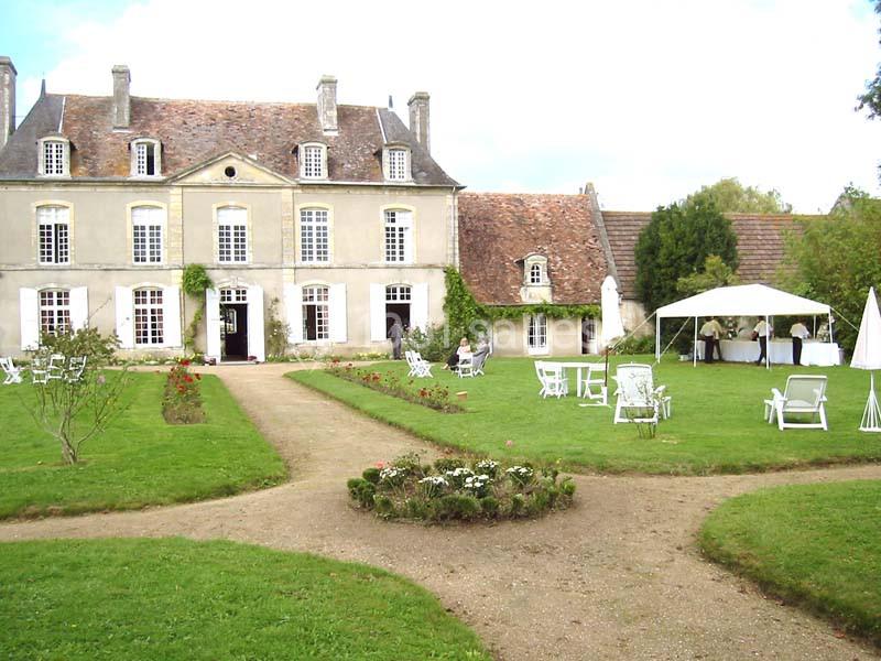 Manoir ancien entouré d'un jardin avec pelouse, fleurs, chaises blanches et une tente installée à droite.