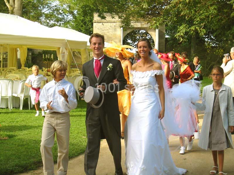 Un couple en tenue de mariage marche dans un jardin accompagné d'enfants et d'invités en arrière-plan.