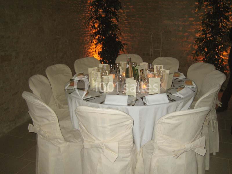 Table ronde dressée pour un dîner, avec nappes blanches, chaises couvertes, menus et bougies, dans une salle en pierre.