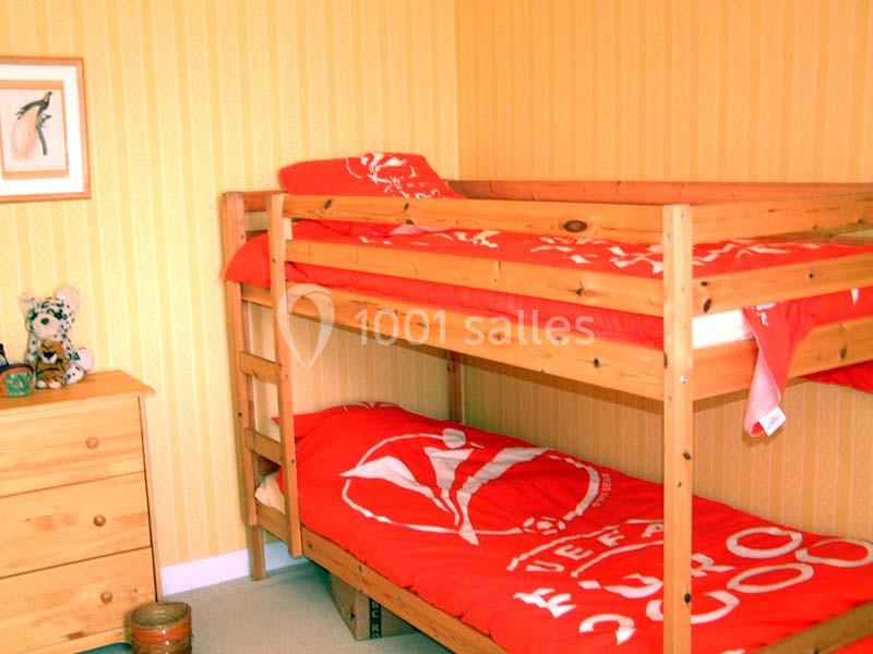 Lit superposé en bois avec draps rouges dans une chambre aux murs jaunes, décorée d'une commode et d'un cadre.