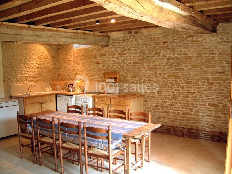 Cuisine rustique avec murs en pierre, poutres apparentes, table en bois entourée de chaises et équipements de cuisine.