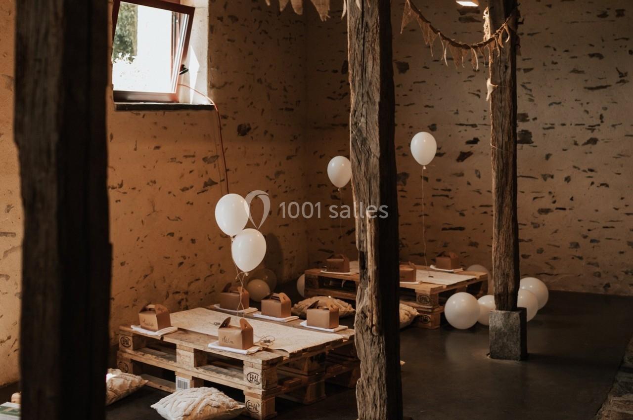 Salle rustique avec tables basses en palettes, coussins au sol et ballons blancs, éclairée par une fenêtre.