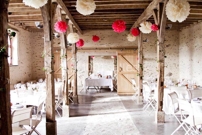 Salle de réception décorée avec des pompons suspendus rouges et blancs, tables dressées et chaises blanches pliantes.