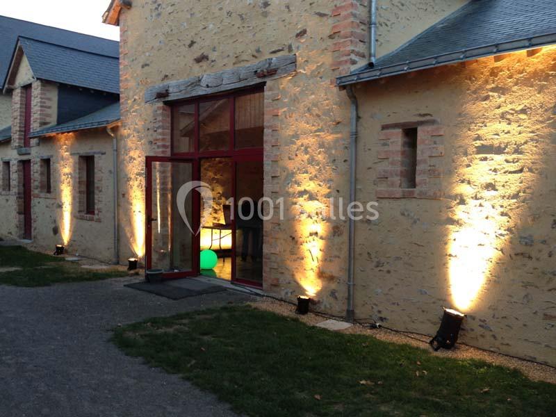 Façade en pierre éclairée par des projecteurs au sol, avec une porte vitrée ouverte donnant sur l'intérieur.