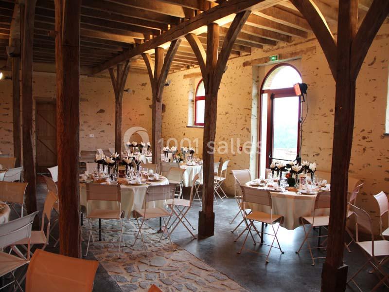 Salle rustique avec poutres apparentes, tables rondes dressées pour un repas, chaises pliantes et lumière naturelle.