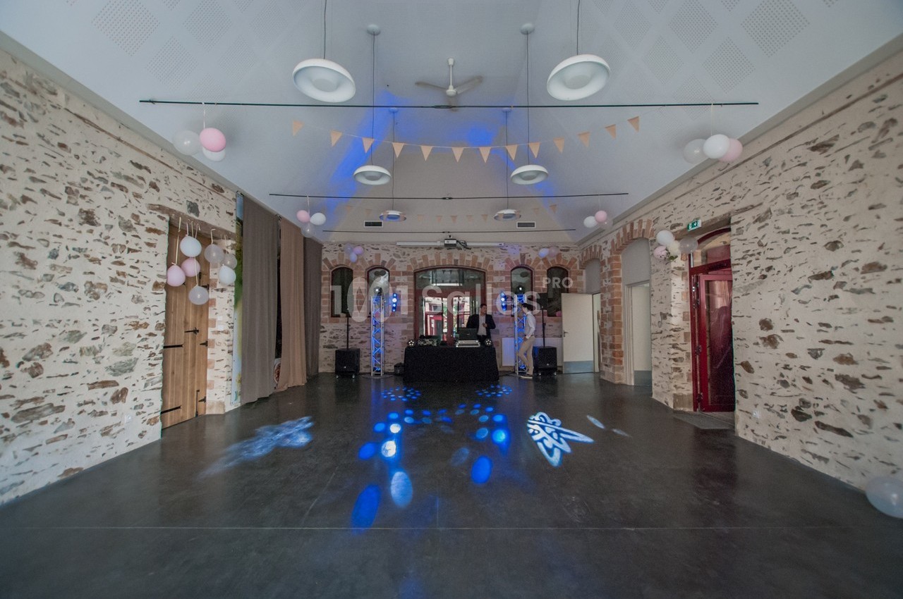 Salle en pierre décorée de guirlandes et ballons, avec une piste de danse éclairée et une table de DJ au centre.