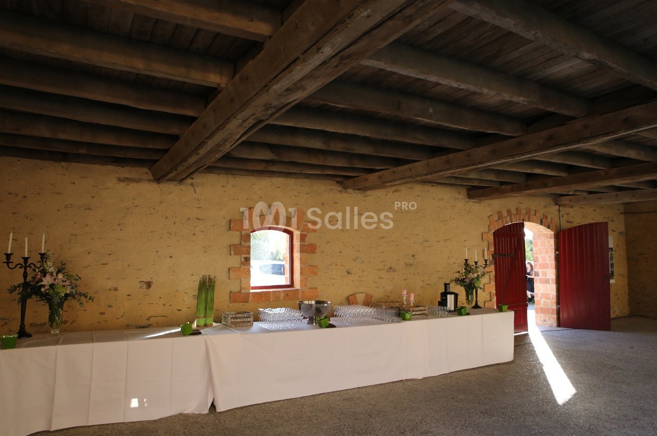 Table dressée avec nappes blanches, verres et décorations florales dans une salle en pierre avec poutres en bois apparentes.