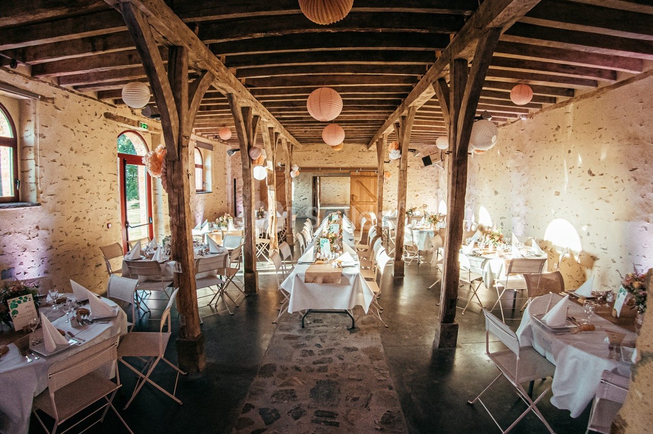 Salle de réception rustique avec tables dressées, chaises blanches et décorations suspendues sous un plafond en bois.