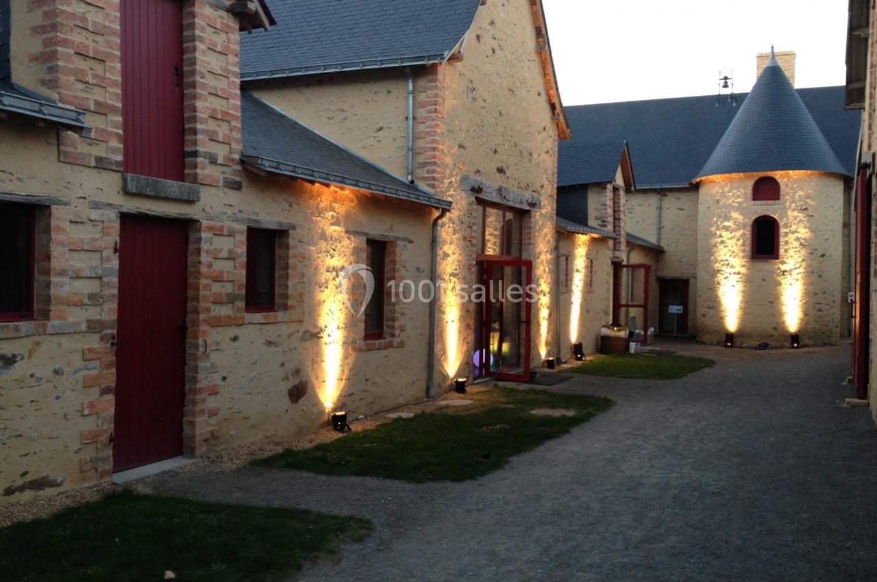 Bâtiments en pierre avec portes rouges, éclairés par des lumières au sol, dans une cour pavée au crépuscule.