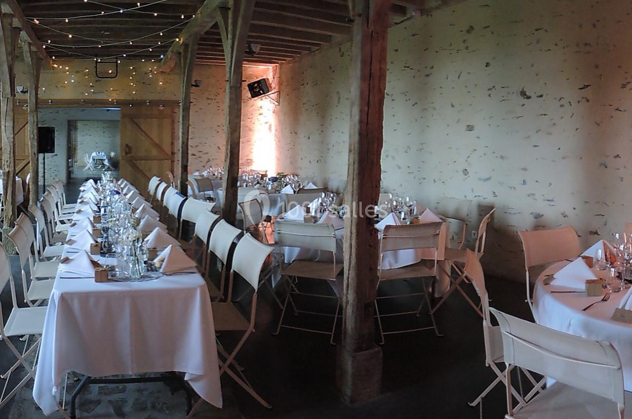 Salle de réception rustique avec tables rondes et rectangulaires dressées pour un événement, éclairage tamisé.