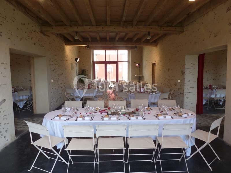 Salle de réception avec tables dressées, nappes blanches, chaises pliantes et lumière naturelle entrant par de grandes baies…