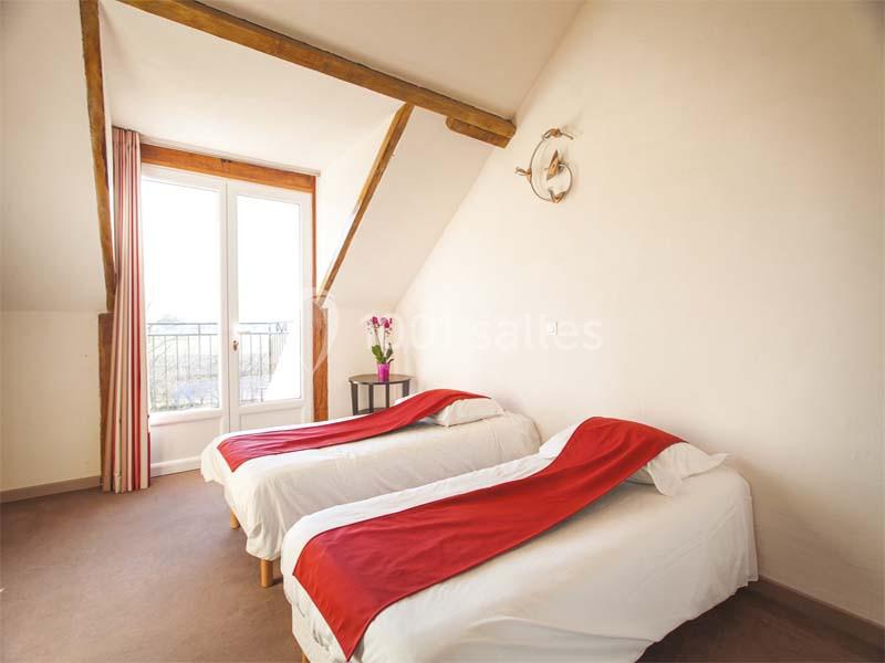 Deux lits simples avec couvre-lits rouges dans une chambre lumineuse avec poutres apparentes et porte-fenêtre donnant sur un…