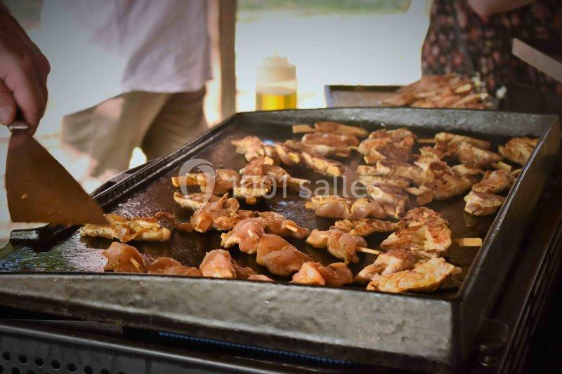 Des brochettes de viande grillent sur une plancha, manipulées à l'aide d'une spatule en extérieur.
