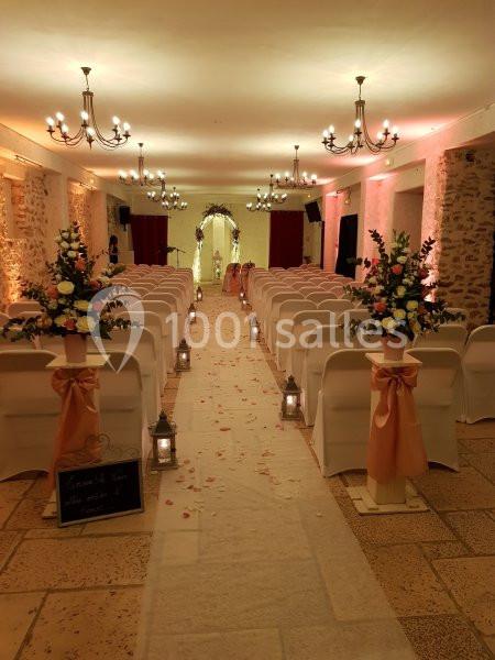 Salle décorée pour une cérémonie, avec rangées de chaises blanches, allée centrale ornée de pétales et arche fleurie au fond.