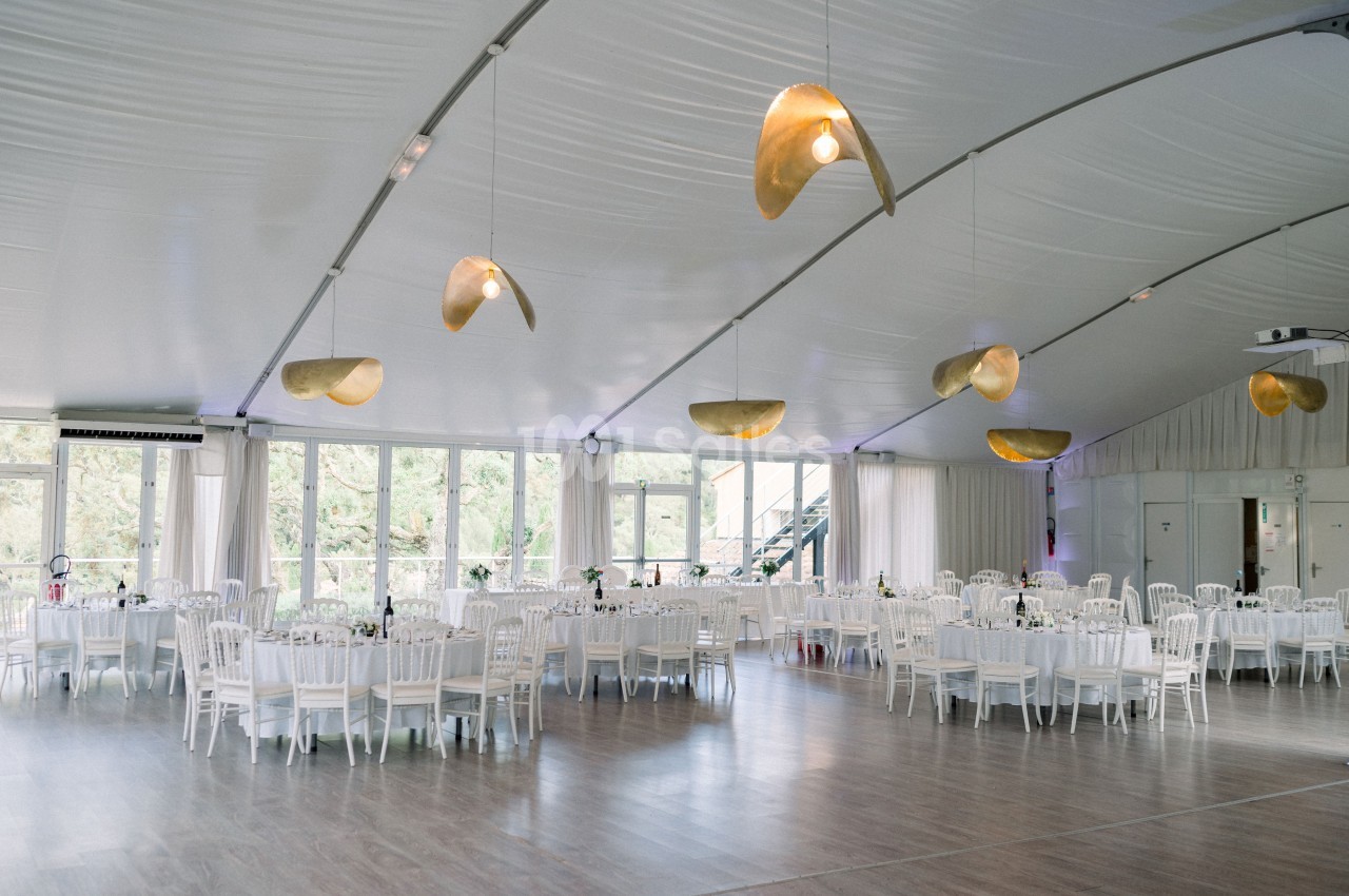 Salle de réception lumineuse avec tables et chaises blanches, décorée de suspensions dorées sous un plafond voûté.