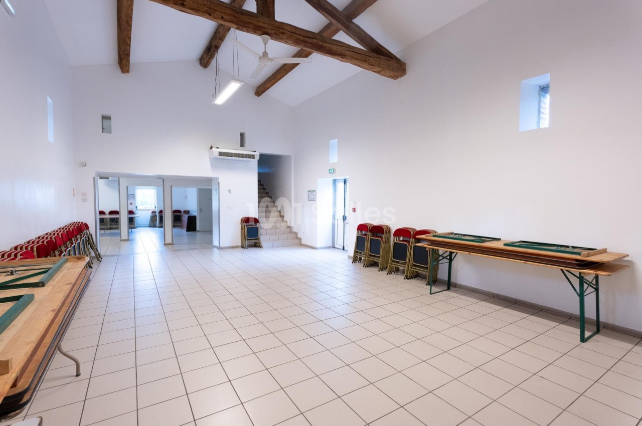 Salle lumineuse avec carrelage clair, poutres apparentes, tables pliantes et chaises empilées contre les murs.
