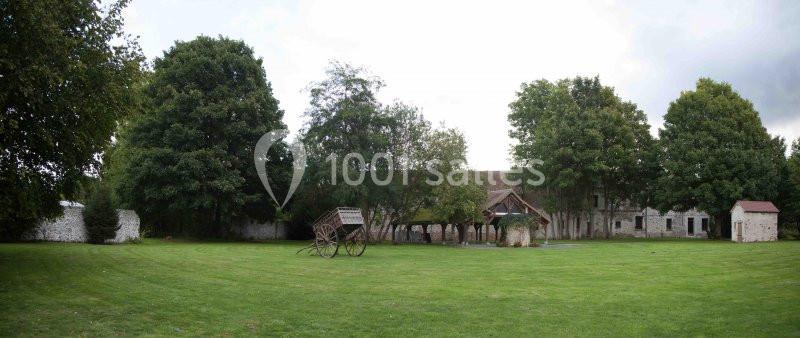 Cour verdoyante avec arbres, charrette en bois, puits et bâtiments anciens en pierre en arrière-plan.