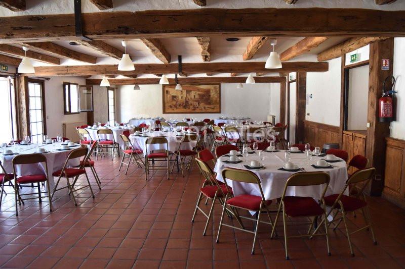 Salle de réception avec tables rondes dressées, chaises rouges, poutres apparentes et sol en carrelage.