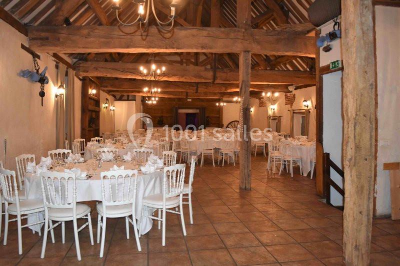 Salle de réception rustique avec poutres apparentes, tables rondes dressées et chaises blanches.