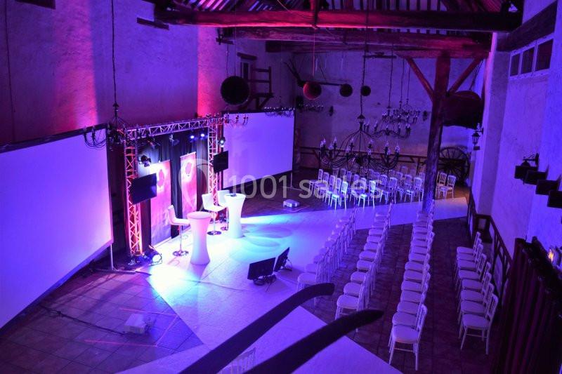Salle de conférence aménagée avec des chaises alignées, un podium éclairé et des écrans projetés dans un décor rustique.
