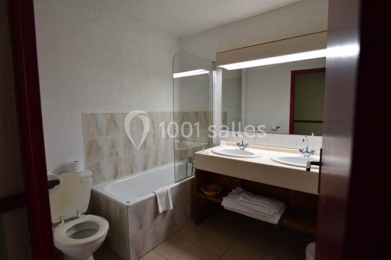 Salle de bain avec baignoire, toilettes, double vasque, miroir éclairé et serviettes pliées sur une étagère.