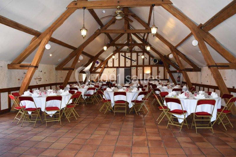 Salle de réception avec poutres apparentes, tables rondes dressées et chaises rouges sur un sol en carrelage.