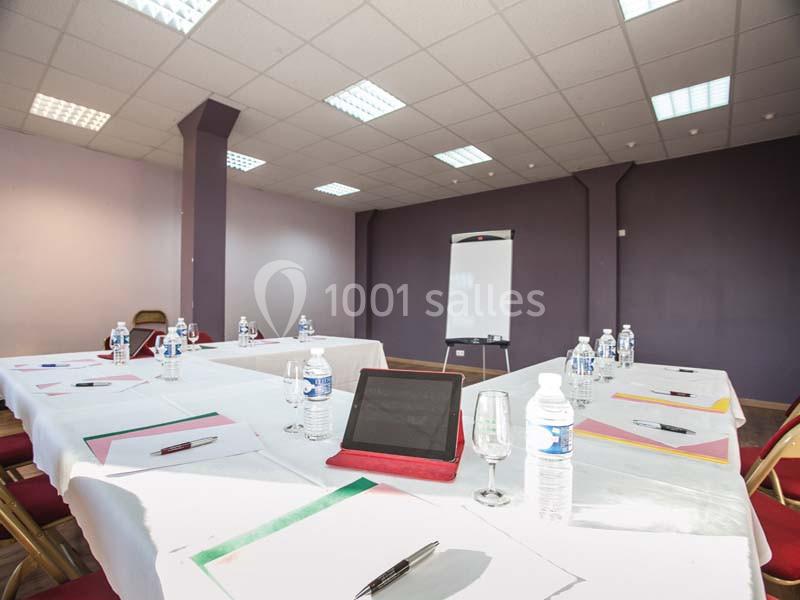 Salle de réunion avec tables disposées en U, documents, bouteilles d'eau, tablette et tableau blanc.
