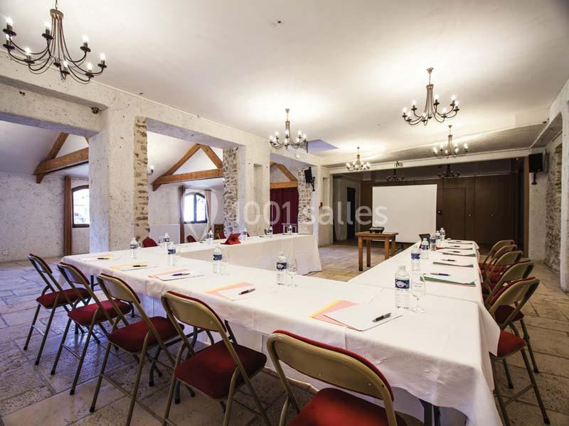 Salle de réunion lumineuse avec tables disposées en U, chaises rouges, projecteur et murs en pierre apparente.