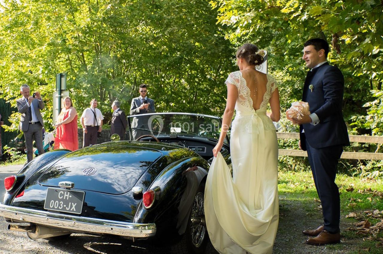 Un couple en tenue de mariage près d'une voiture ancienne noire, entouré d'invités dans un cadre verdoyant.