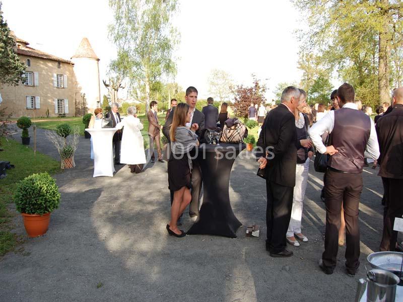 Groupe de personnes discutant debout autour de tables hautes en extérieur, près d'un bâtiment ancien et d'arbres.