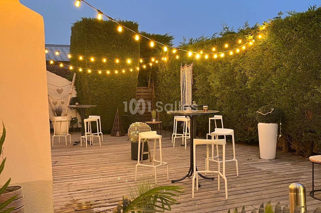Terrasse en bois éclairée par des guirlandes lumineuses, avec tables hautes, tabourets et haies en arrière-plan.