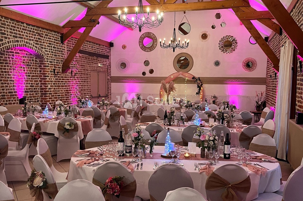 Salle de réception décorée pour un événement, avec tables rondes, chaises ornées et éclairage rose.