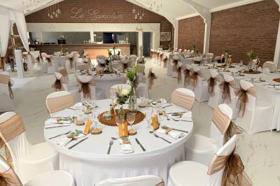 Miniature Location salle Paillencourt (Nord) - Le Canotier #3 Table en bois décorée de bougies, fleurs et feuillage, entourée de chaises, sous une arche végétale dans une salle lumineuse.