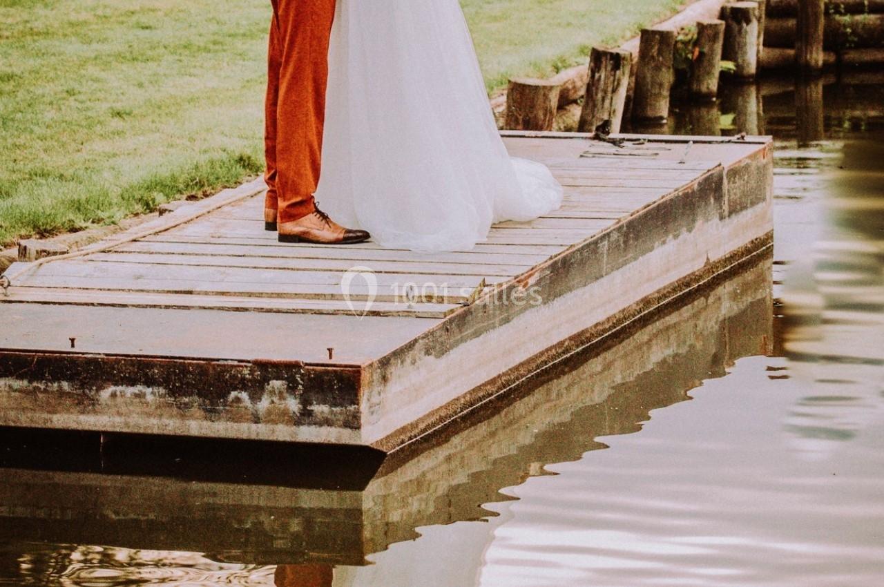 Un couple s'embrasse sur un ponton en bois au bord d'un plan d'eau, entouré de verdure.