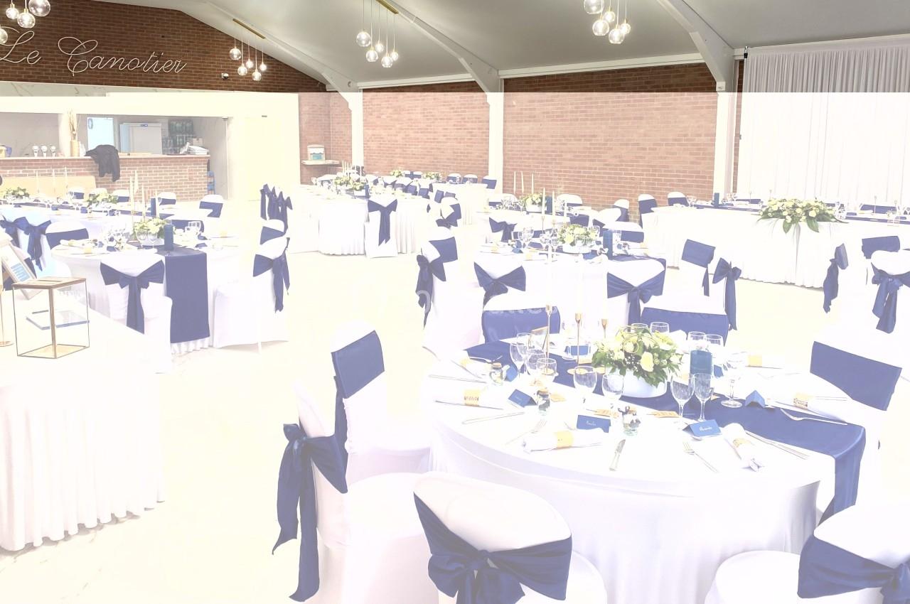 Salle de réception décorée avec des nappes blanches et des rubans bleus sur les chaises, tables dressées pour un événement.
