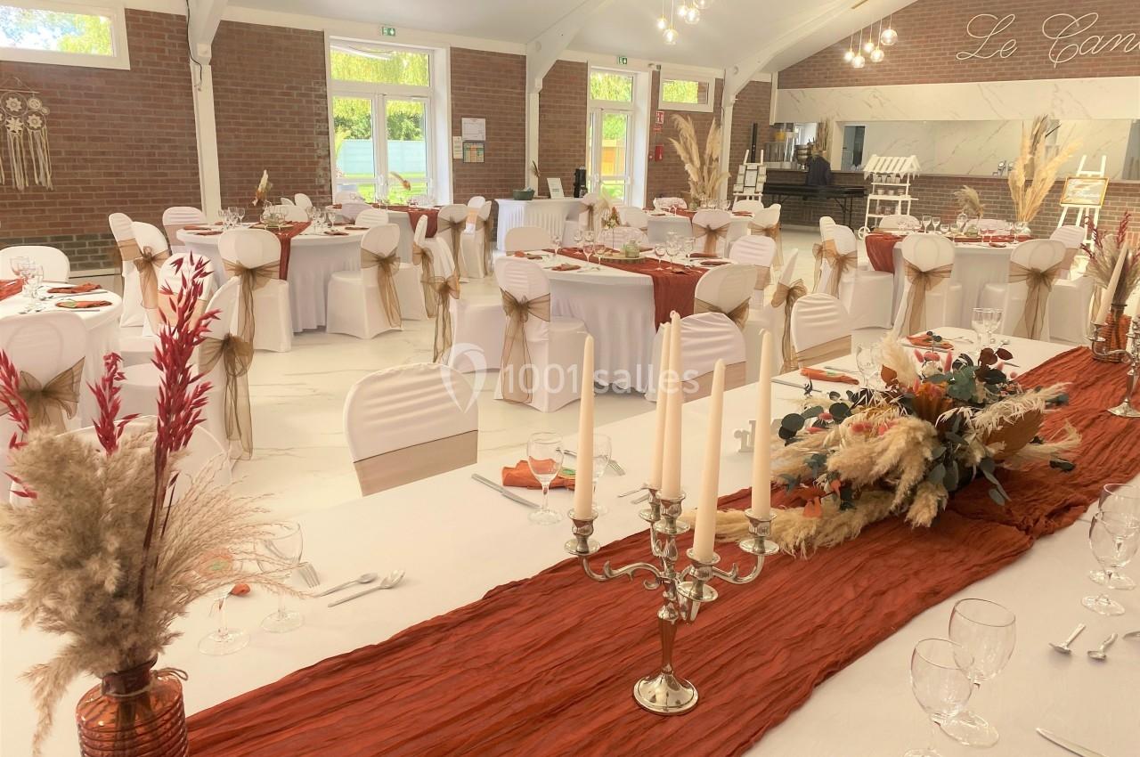 Salle de réception décorée avec des tables dressées, nappes blanches, chemins de table rouille et compositions florales.
