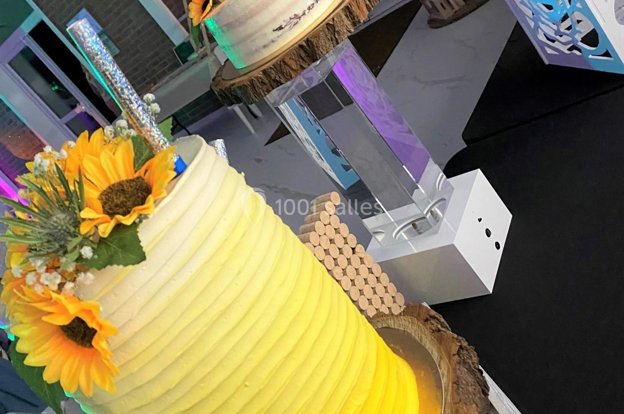 Gâteau cylindrique jaune décoré de tournesols et d'éléments brillants, posé sur une base en bois dans une salle éclairée.