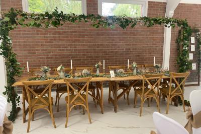 Miniature Location salle Paillencourt (Nord) - Le Canotier #43 Table en bois décorée de bougies, fleurs et feuillage, entourée de chaises, sous une arche végétale dans une salle lumineuse.
