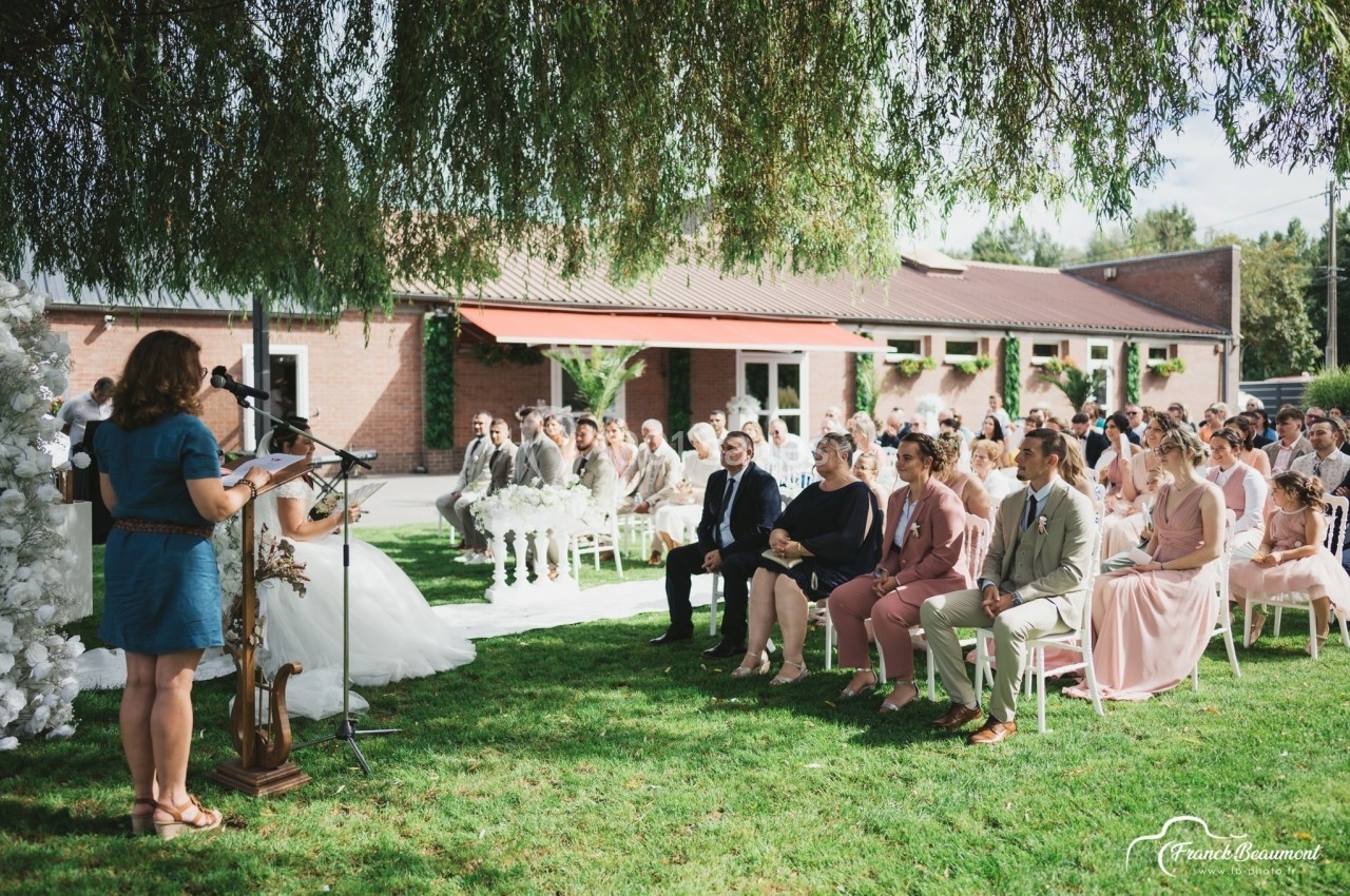 Cérémonie en plein air avec des invités assis sous un arbre, face à une mariée et une intervenante au micro.