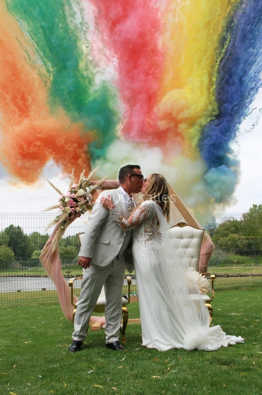 Un couple en tenue de mariage s'embrasse devant une arche florale, avec des fumées colorées en arrière-plan.