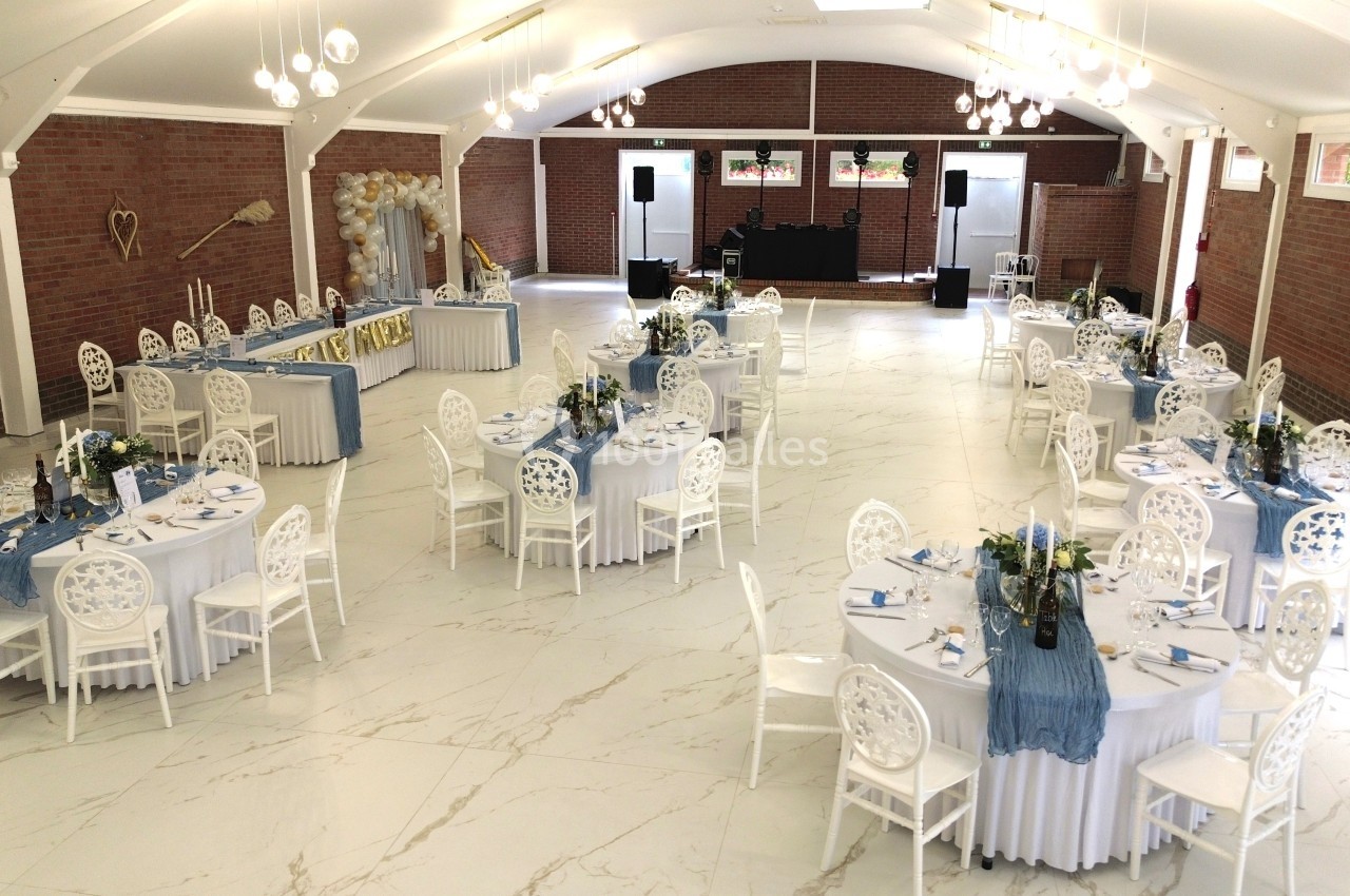 Salle de réception lumineuse avec tables rondes décorées de nappes bleues et blanches, prête pour un événement.