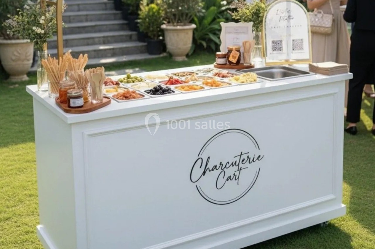 Chariot blanc avec un assortiment de charcuteries, fromages et condiments, disposé sur une pelouse extérieure.