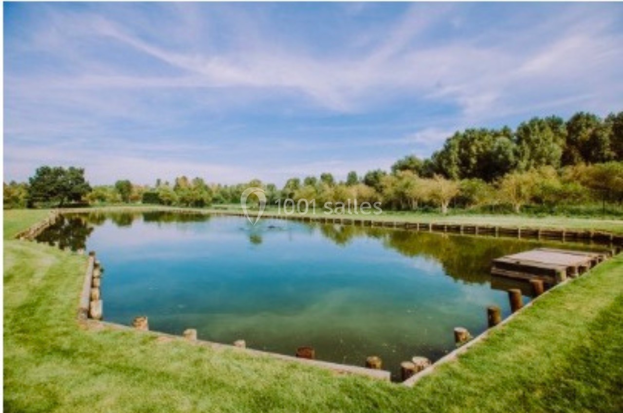 Étang entouré de pelouse et d'arbres sous un ciel bleu avec quelques nuages.