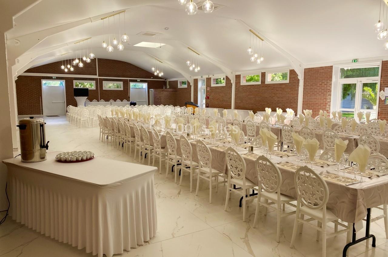 Salle de réception lumineuse avec de longues tables dressées, chaises blanches et décorations élégantes.