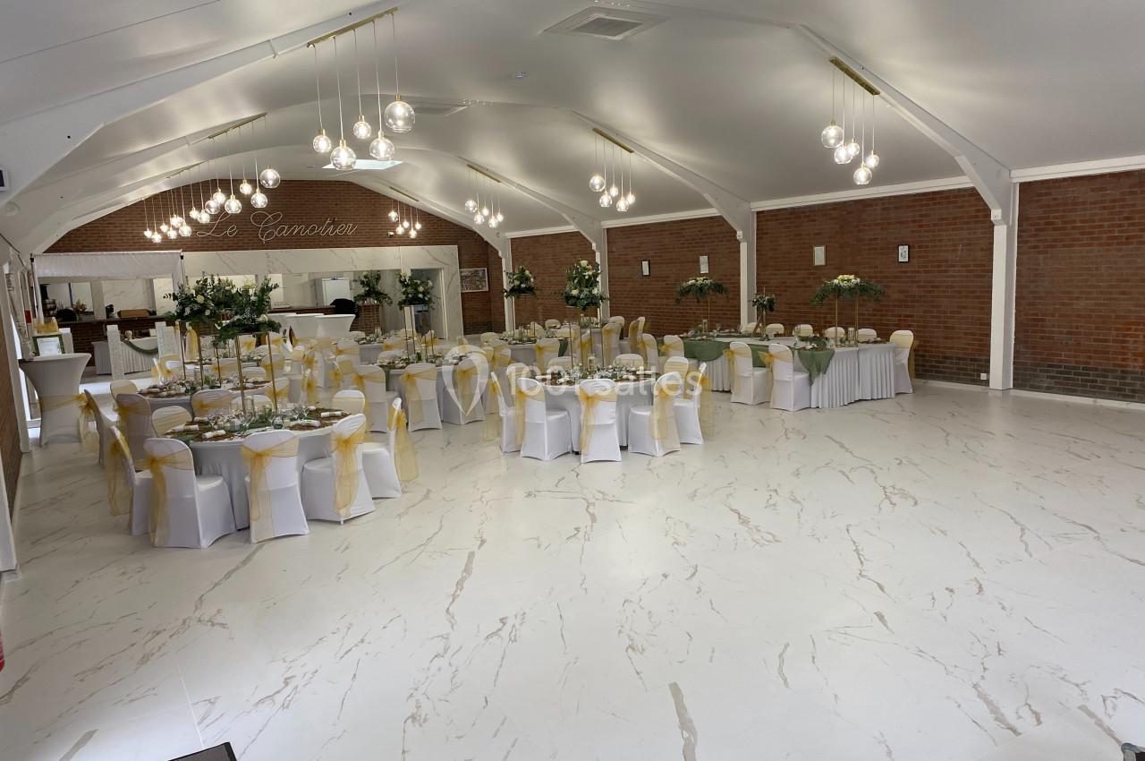 Salle de réception lumineuse avec tables rondes décorées de nappes blanches, chaises ornées de rubans jaunes et centres de…